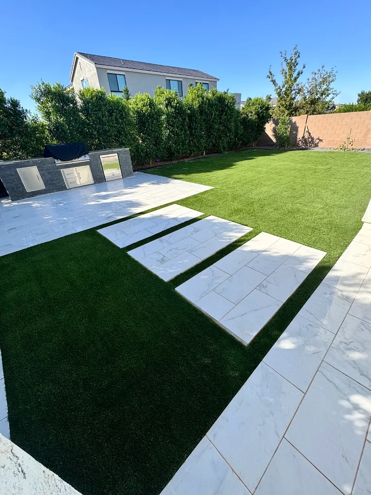 A modern backyard landscape featuring a vibrant artificial grass lawn divided by large white marble-look stepping stone pavers, accompanied by a matching paved patio area and an outdoor kitchen counter under a clear blue sky.