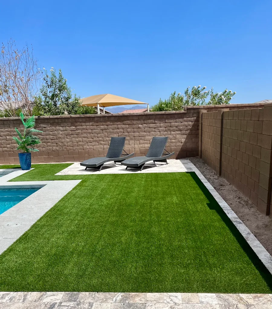 A pristine backyard space featuring a rectangular artificial grass lawn bordered by stone pavers, two dark lounge chairs positioned on a patio area against a brown block wall, and a partial view of a swimming pool under a bright blue sky.