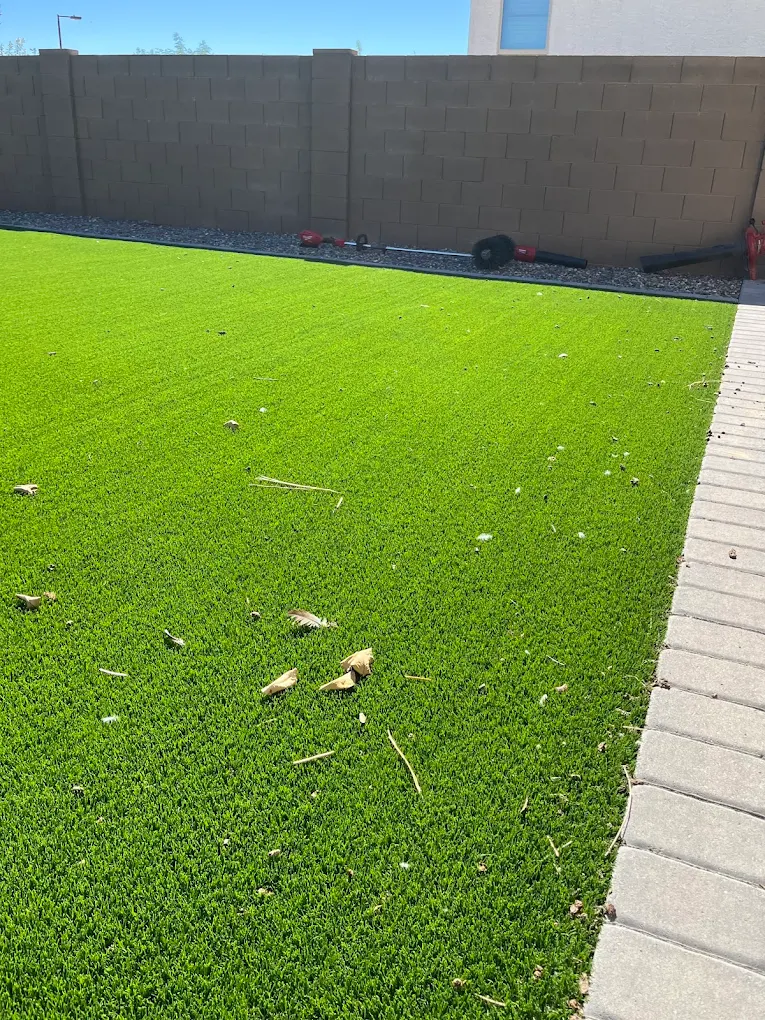 A vibrant green artificial grass lawn scattered with dry leaves and debris, bordered by a light-colored paver walkway on the right and a tall brown block wall in the background under a clear sky.
