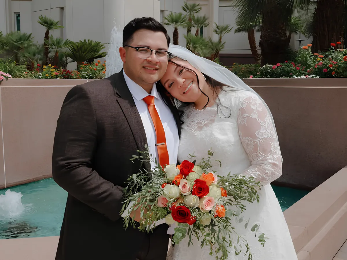 A happy newlywed couple, the bride holding a vibrant bouquet, standing in a sunny garden near a fountain.