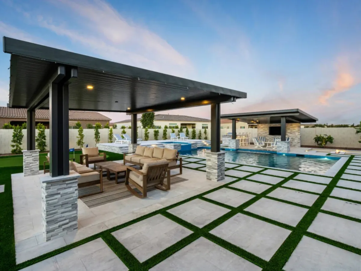 A luxurious outdoor space with a large pool, shaded seating area with a sofa, and a stone bar under a modern black patio cover at dusk.
