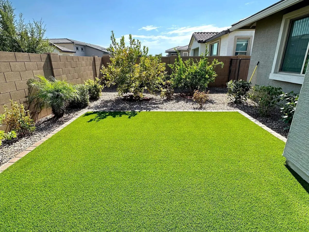 A modern backyard features bright green artificial turf surrounded by a gravel border with desert-friendly plants and a block wall.