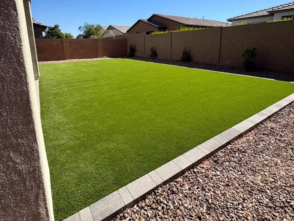 A front yard view of a single-story stucco home with arched entryways, featuring a neat rectangular artificial turf lawn bordered by pavers and gravel landscaping under an overcast sky.