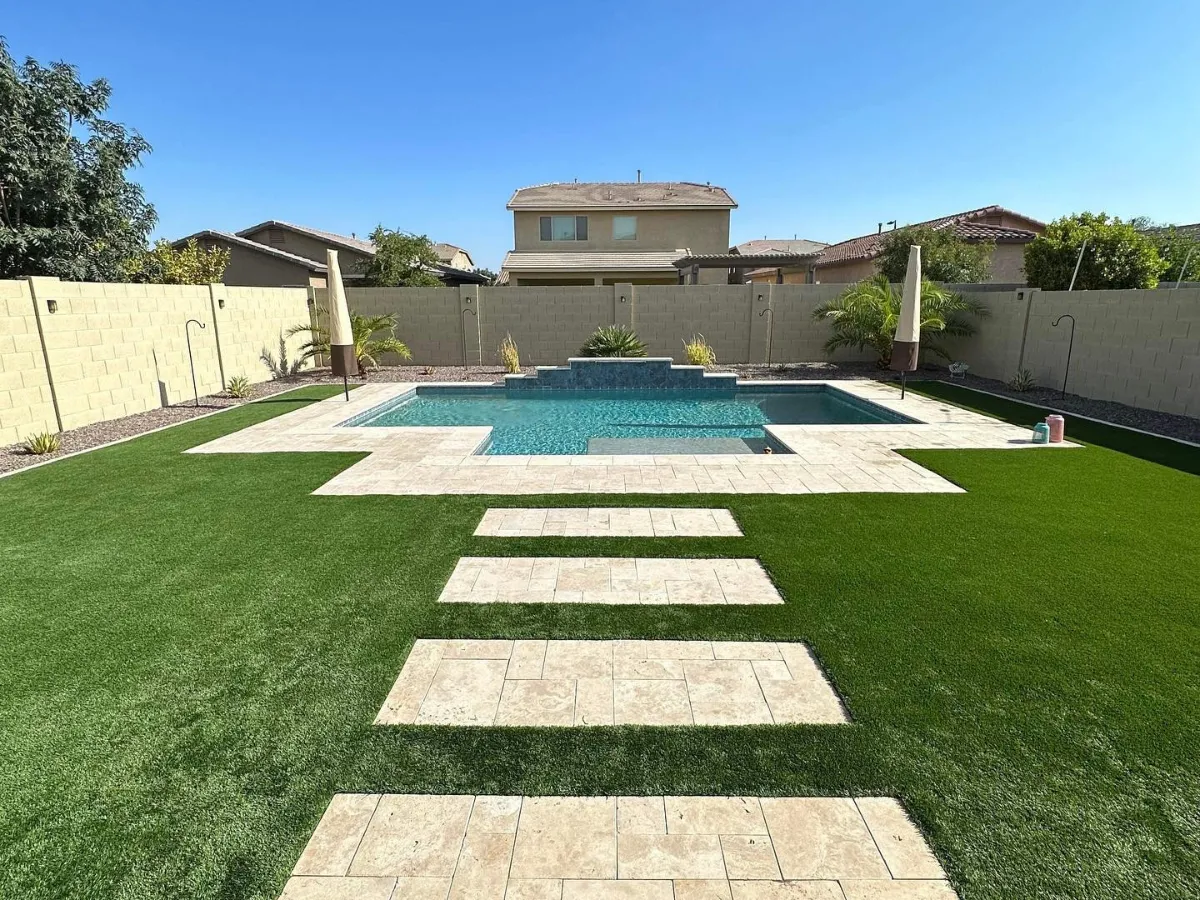 A modern backyard landscape featuring large light-colored stone pavers set in a grid pattern with artificial turf ribbons, alongside a swimming pool on the right and a covered patio area on the left under a bright blue sky.