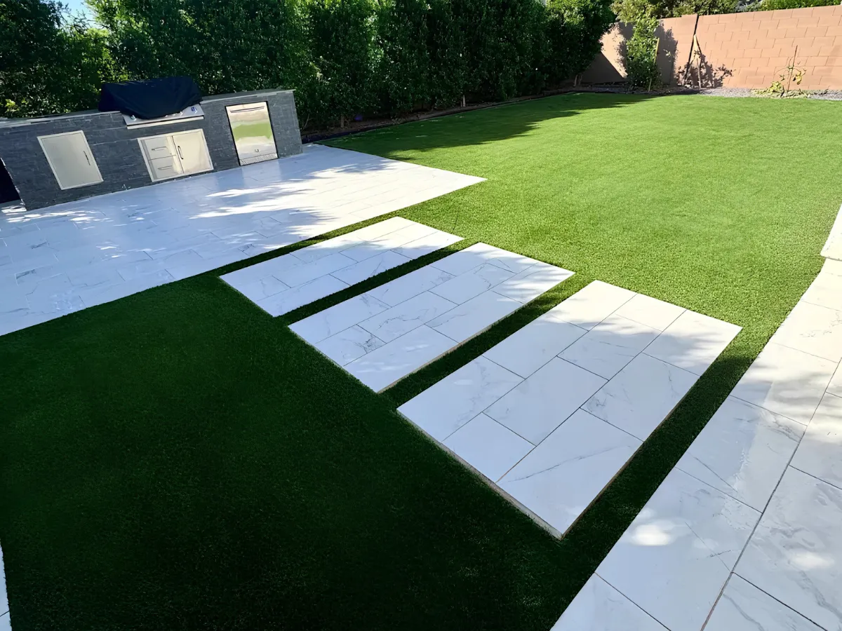 Modern backyard with artificial turf, light tile patio, stone outdoor kitchen, and stepping stone path.