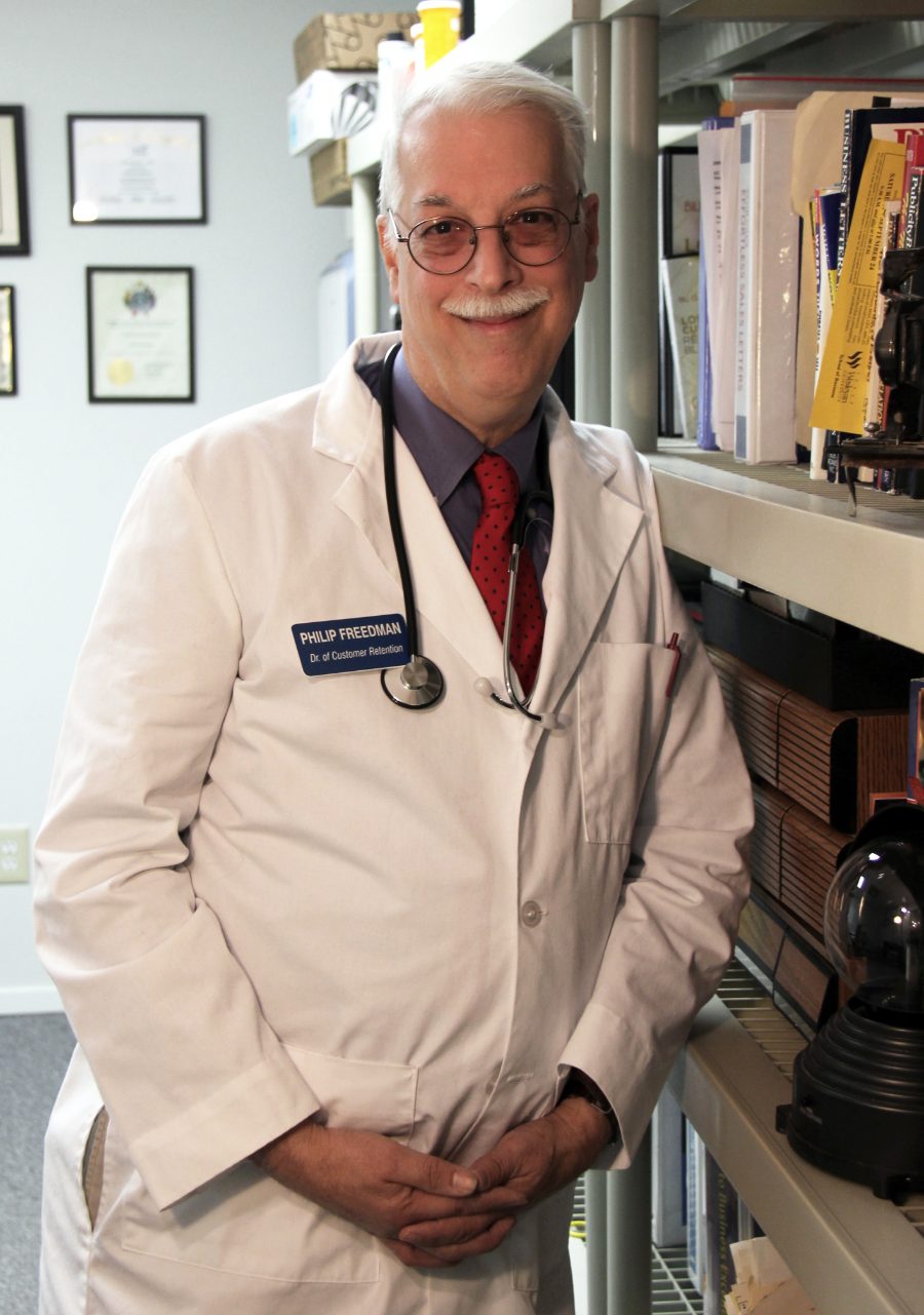 Philip Freedman, owner of My Marketing Cure, wearing a white lab coat and stethoscope with a badge reading Dr. of Customer Retention, in his Plano, Texas office