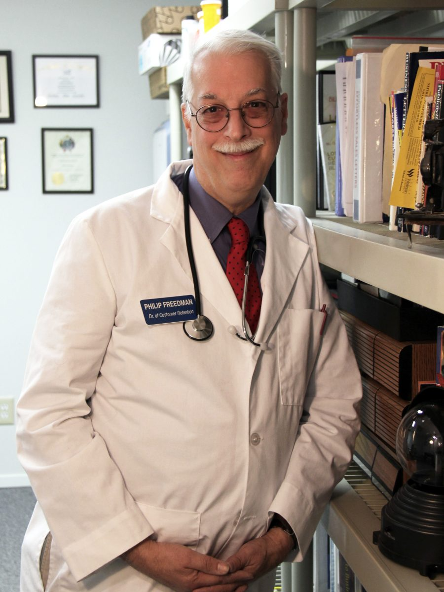 Philip Freedman, owner of My Marketing Cure, standing in his Plano office in a white lab coat with a stethoscope and a Dr of Customer Retention name badge