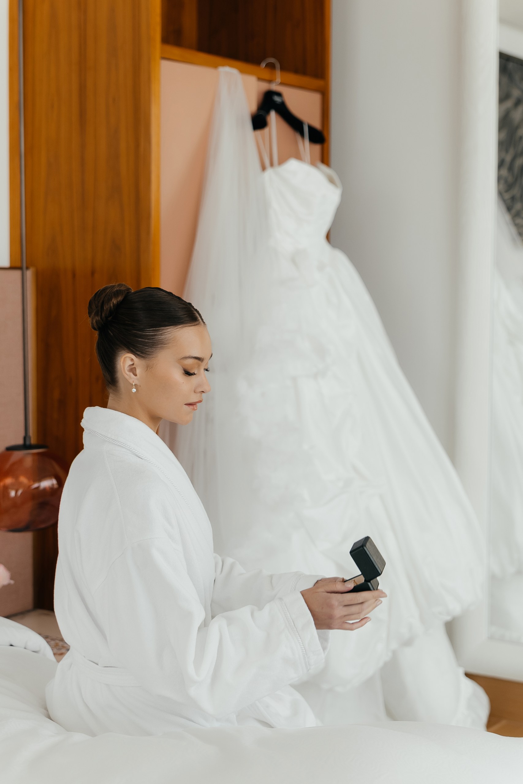 Bride in a white robe holding an open ring box, with her wedding dress hanging behind her at The Lana, Dubai.