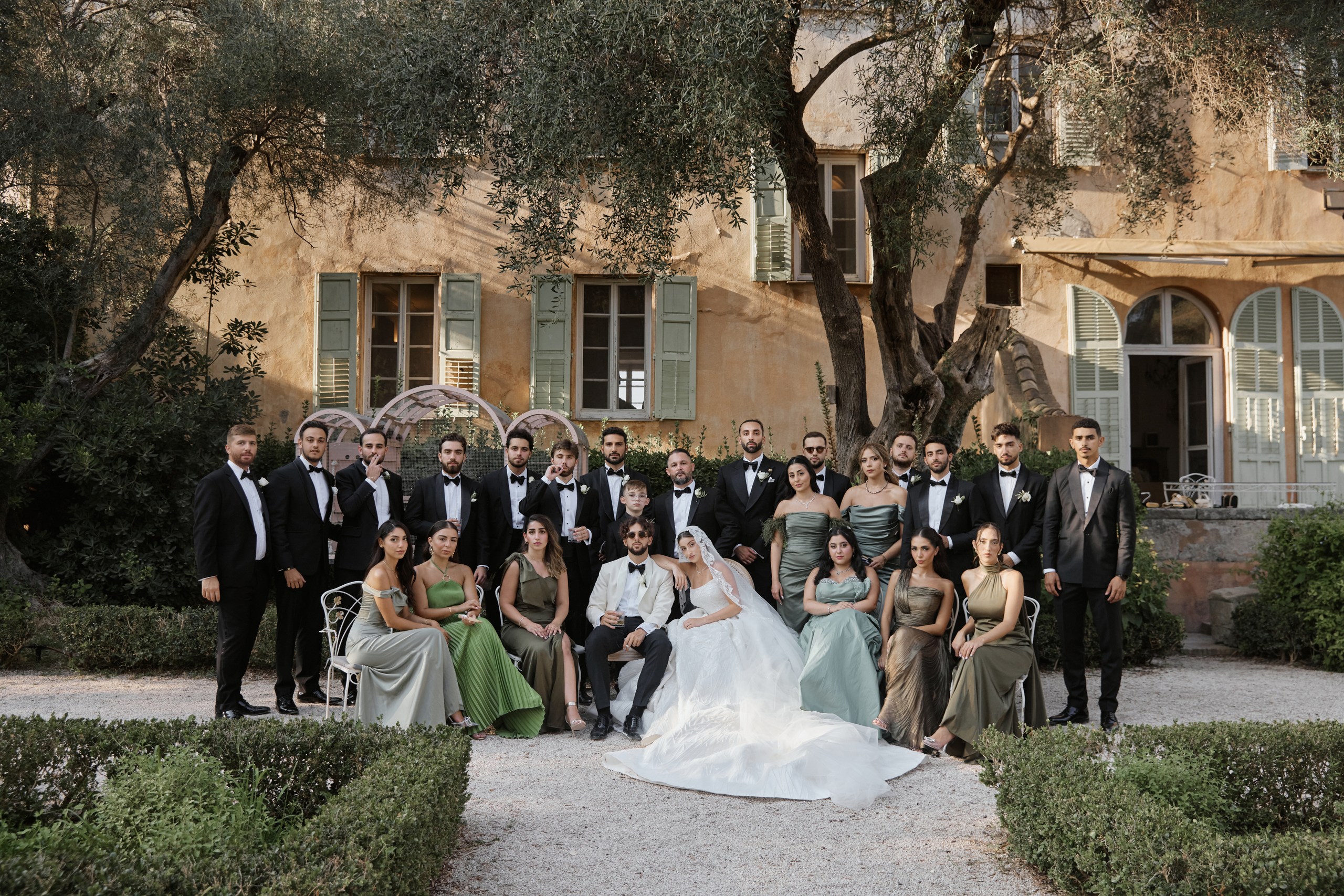 Bride and groom posing with wedding guests beneath olive trees in front of a villa with pale green shutters.