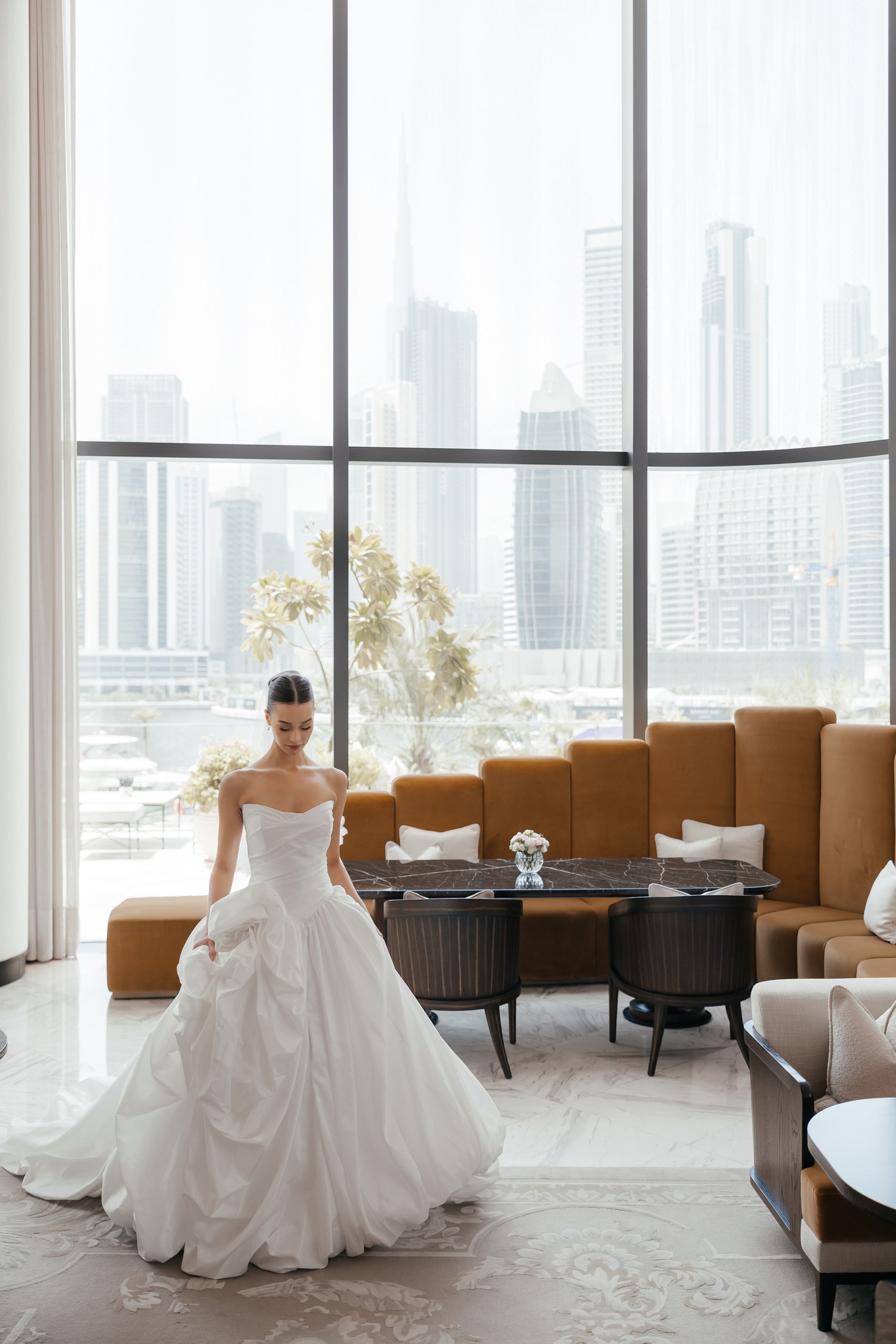 Bride in a white wedding gown beside floor-to-ceiling windows overlooking the Dubai skyline at The Lana hotel.