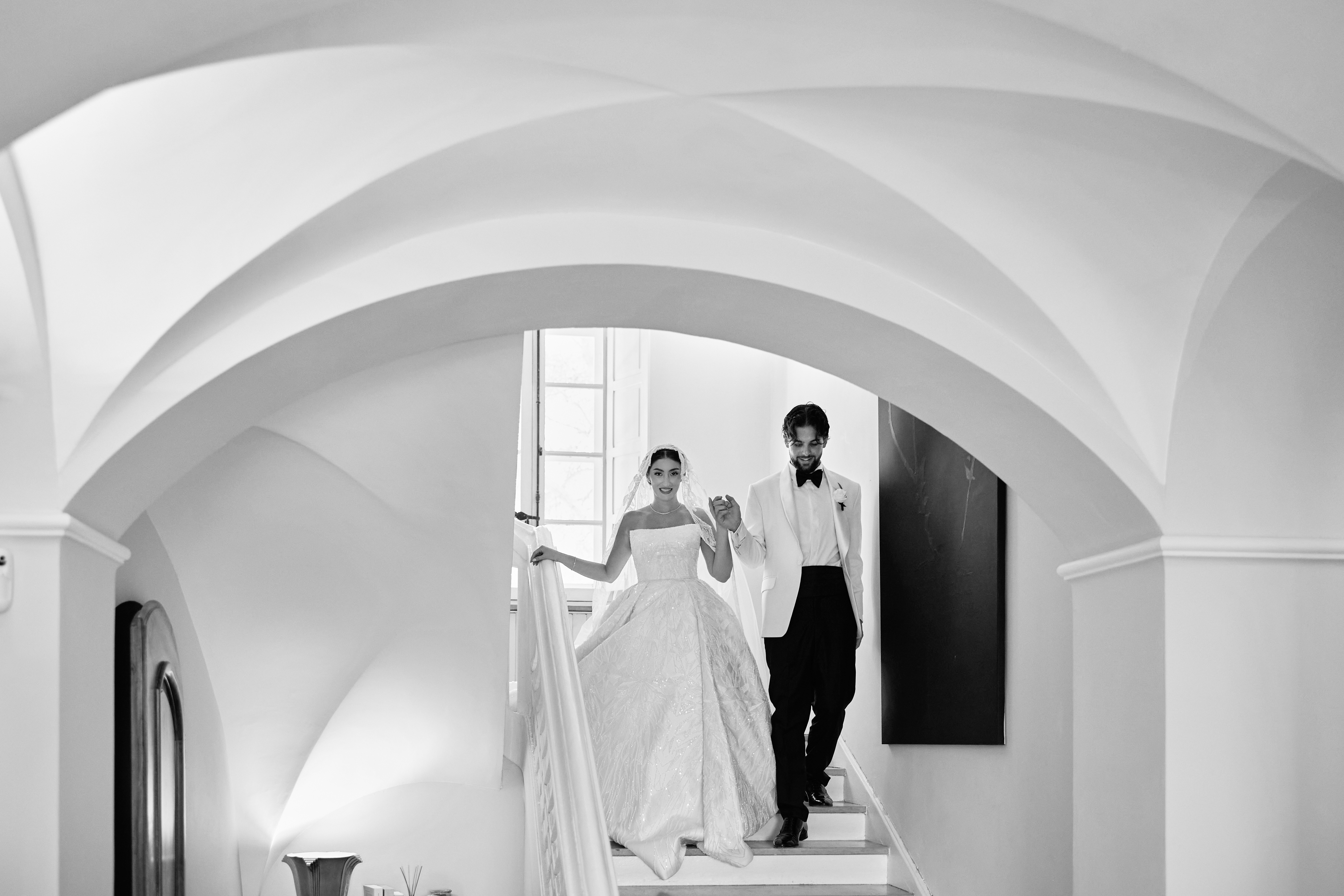 Black and white photograph of a bride and groom holding hands as they descend a staircase beneath vaulted arches.