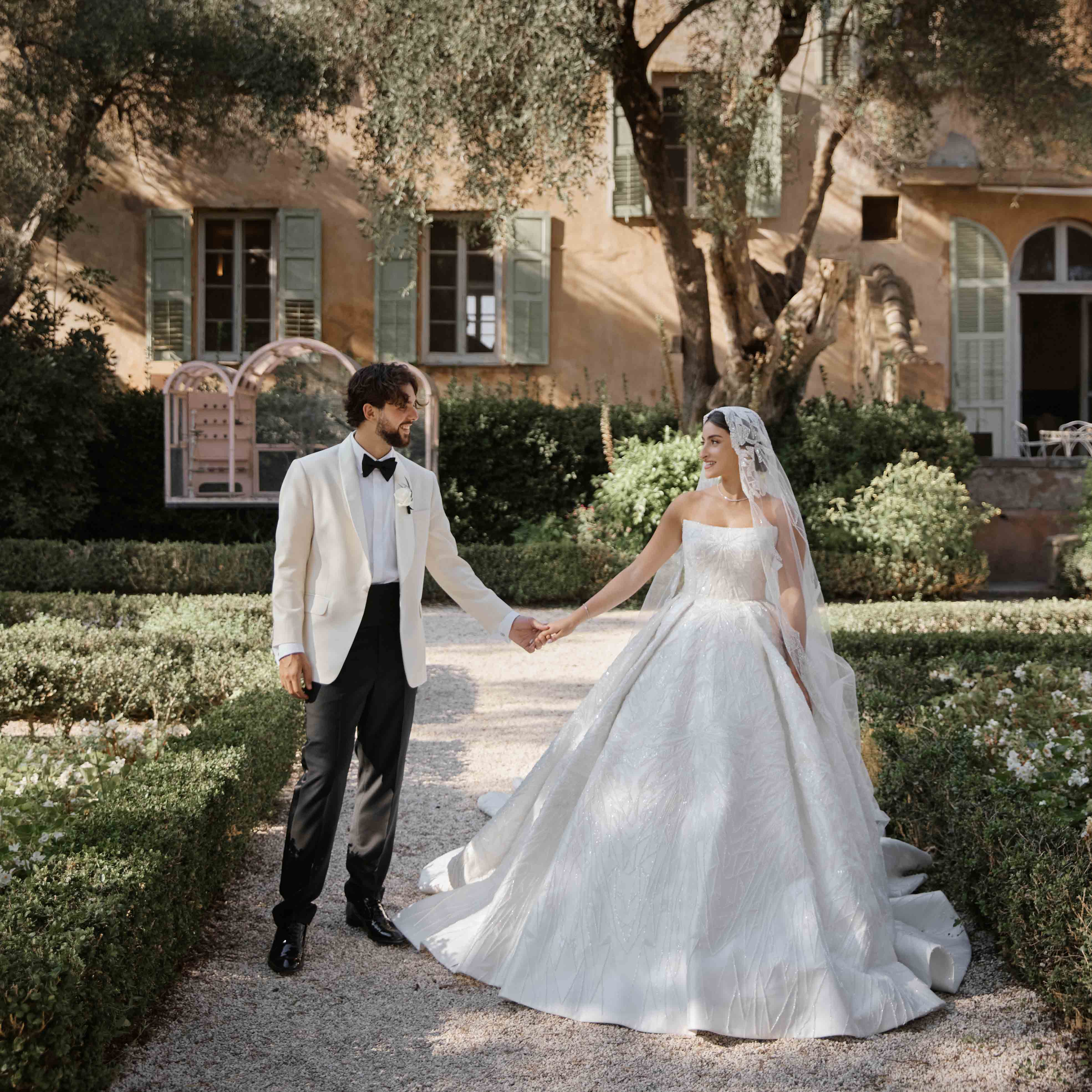 Bride in a white gown and lace veil holding hands with her groom in a white tuxedo jacket in a sunlit villa garden.