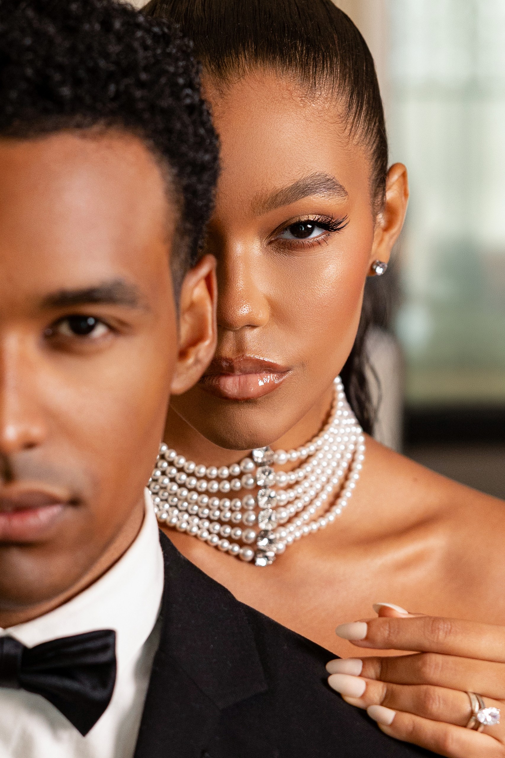 Close-up of a bride wearing a layered pearl necklace, resting her hand on the shoulder of her groom in a black tuxedo.