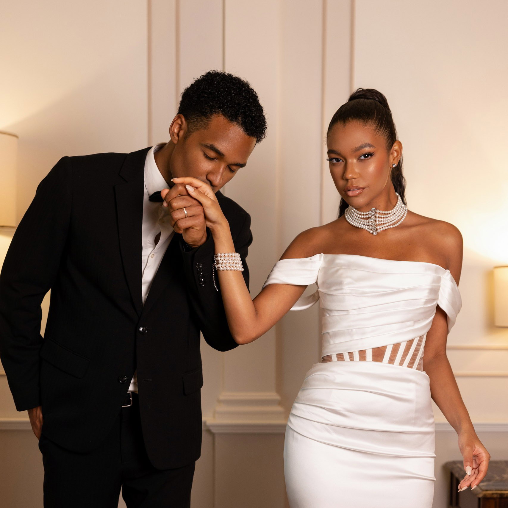 Groom in a black tuxedo kissing the hand of his bride, who wears an off-the-shoulder white gown and layered pearl jewellery.