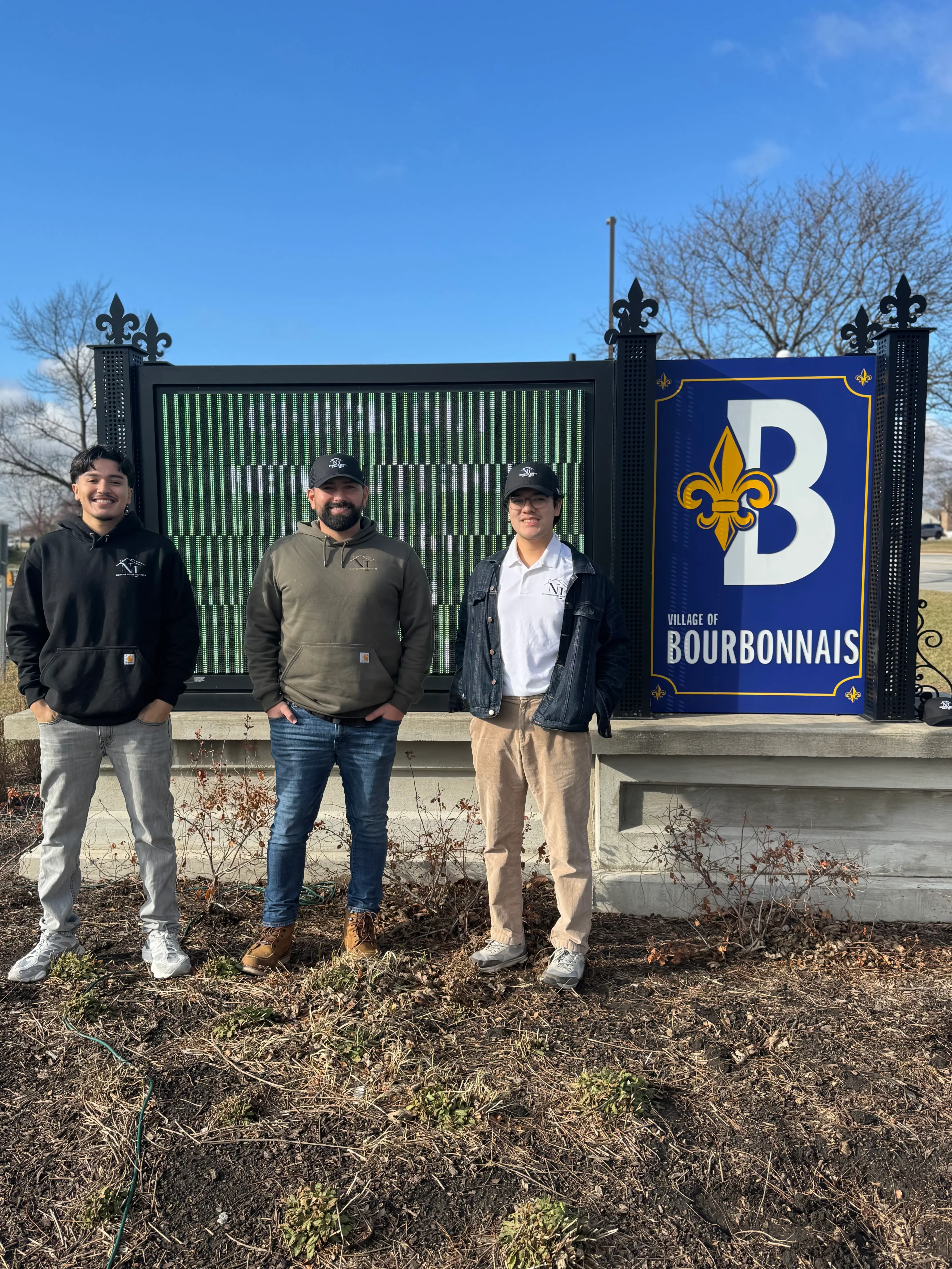 Three N&I Roofing team members in company apparel standing by the Village of Bourbonnais sign in Illinois