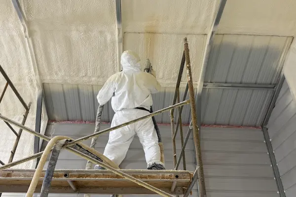 Professional applying spray foam insulation to metal building ceiling for moisture control and energy efficiency.
