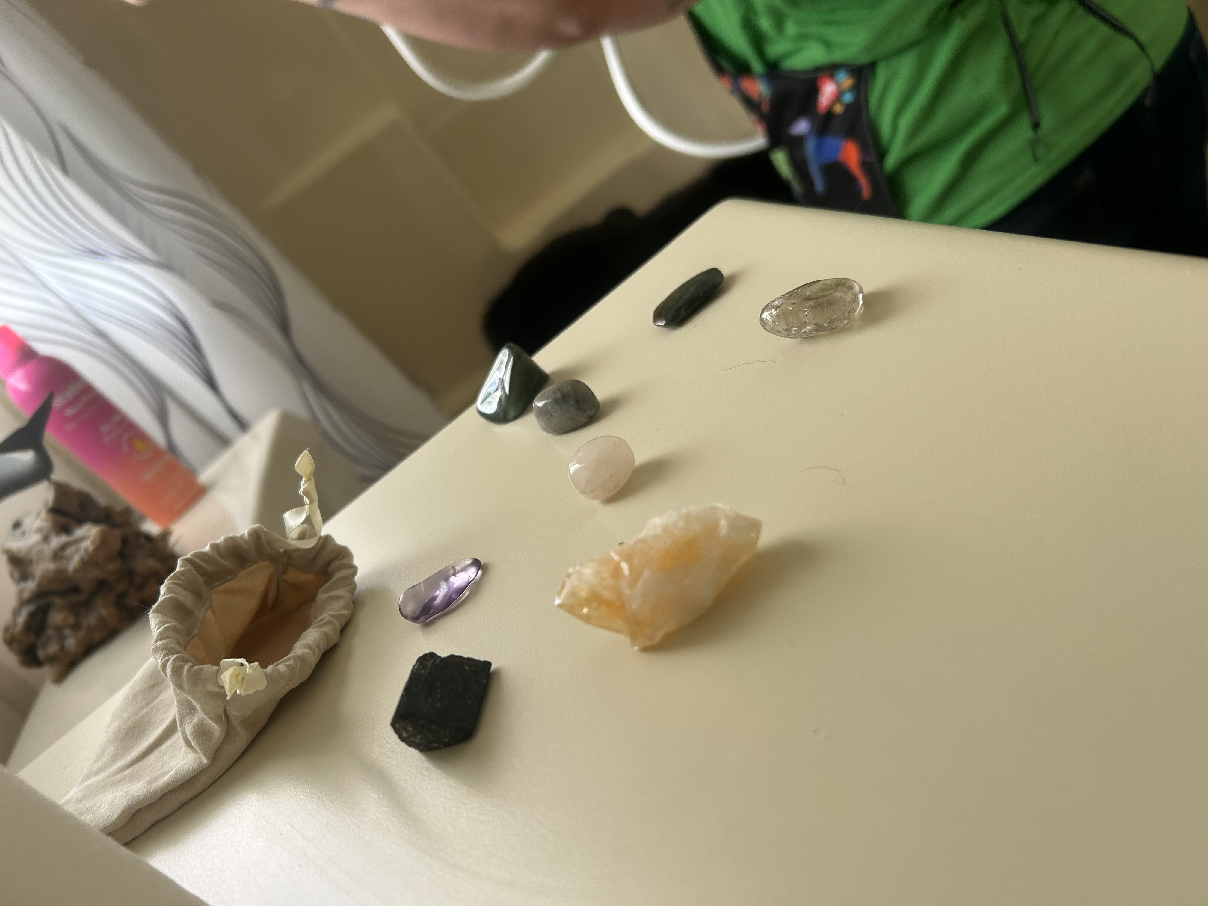 Small collection of polished and raw crystals arranged on a light-colored tabletop, including clear, purple, green, and amber-toned stones, displayed beside a fabric drawstring pouch.