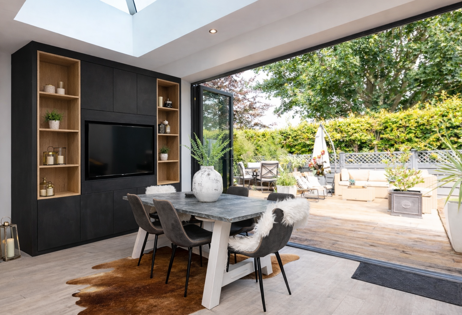 Modern open-plan dining area with a fitted media wall, shelving and TV beside large bifold doors leading to a garden patio