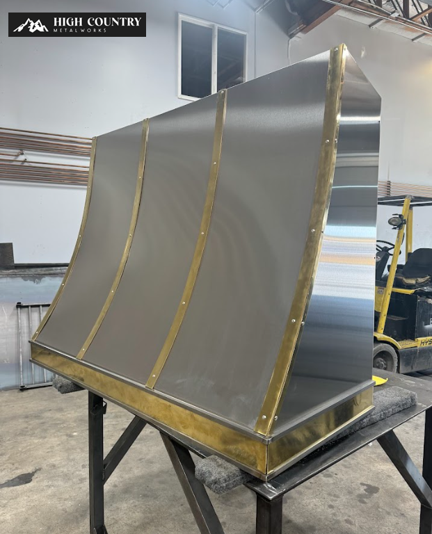 Industrial kitchen hood with a sleek metallic finish and brass accents displayed on a workbench in a workshop.
