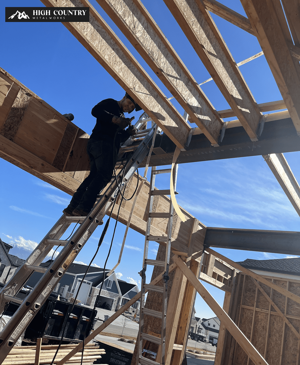 Construction worker on a ladder drilling into wooden beams of a house under blue skies in Provo, UT.