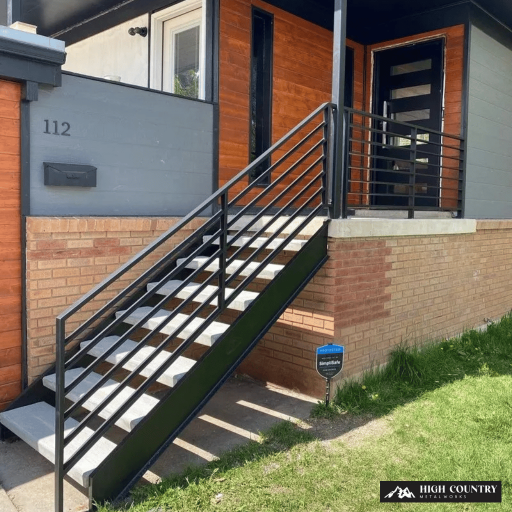 Modern wood and brick house entrance with black wrought iron railing staircase leading to a dark front door on a sunny day.