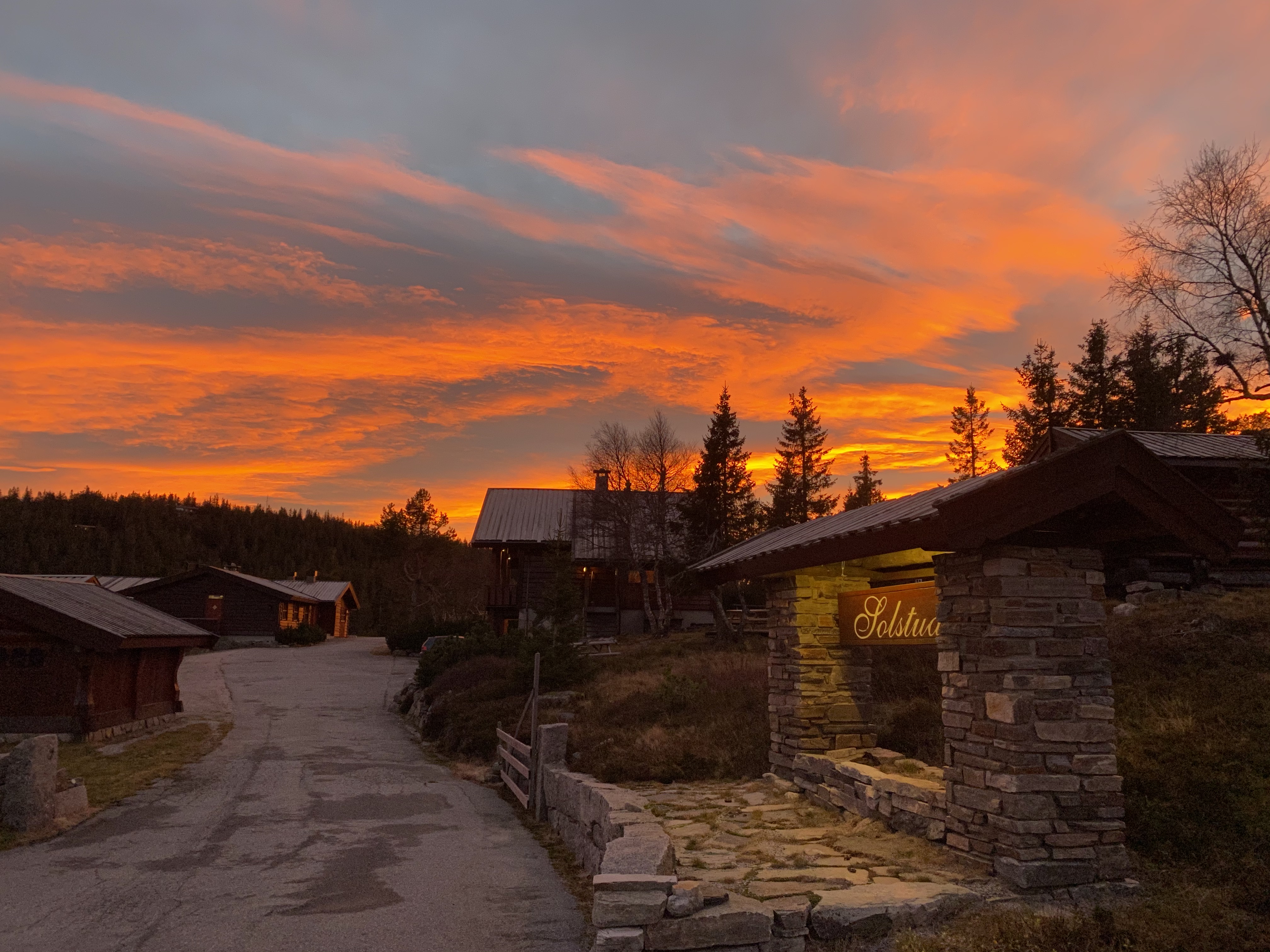 Solstua Lifjell ved solnedgang — steinmur med Solstua-skilt, tømmerhytter og en dramatisk oransje og rosa himmel