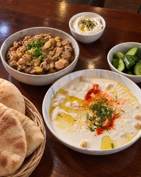 Fatteh in a white bowl beside a plate of foul, with Lebanese bread and cucumbers