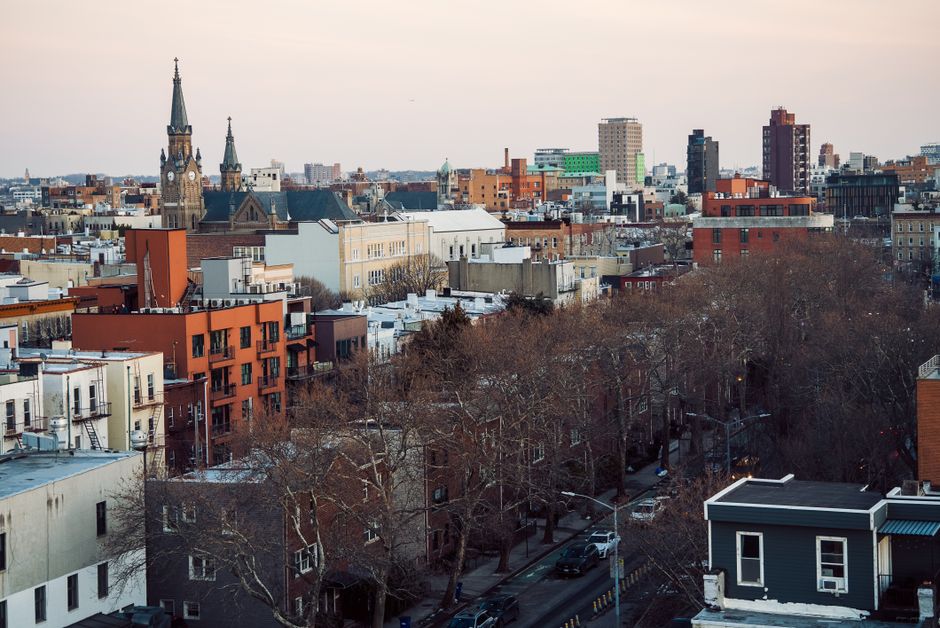 Elevated view over Brooklyn rooftops and bare trees at dusk, with a church spire and low brick buildings.