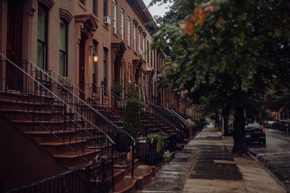 Row of brownstone stoops along a tree-lined Brooklyn sidewalk after rain.
