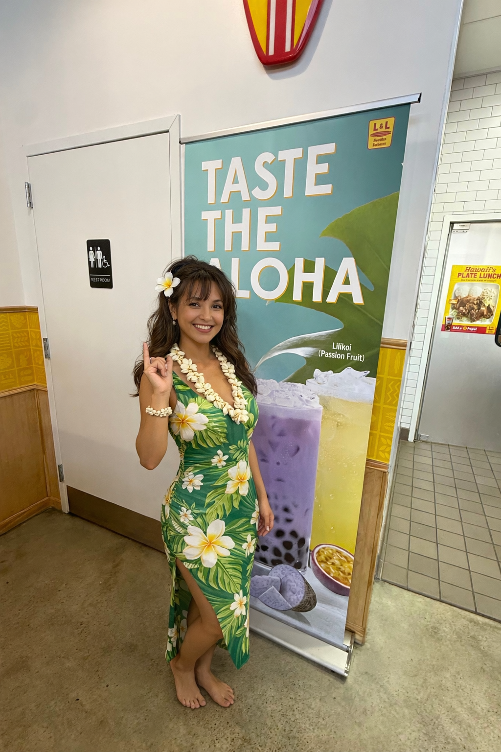 Younger Hawaiian woman in a green tropical muumuu making a shaka sign inside L&L Hawaiian Barbecue in Hamilton Mill, Dacula GA, standing next to a “Taste the Aloha” promotional banner