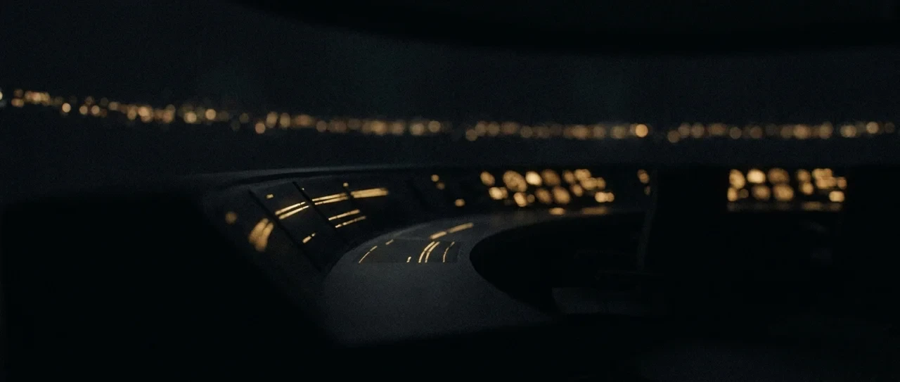 A darkened flight deck at night, instrument panels lit in amber with city lights on the horizon beyond the glass