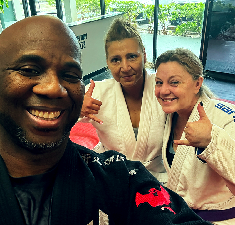 A martial arts instructor taking a selfie on the mats with two smiling female BJJ students throwing shaka signs in white gis. A martial arts instructor taking a selfie on the mats with two smiling female BJJ students throwing shaka signs in white gis.