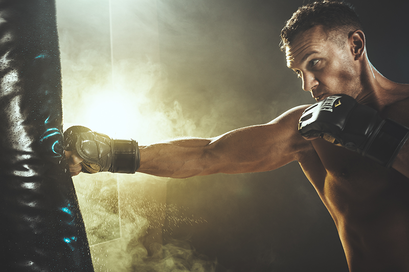 Male athlete in black MMA gloves striking a heavy bag with a powerful jab in a moody, backlit training facility. Male athlete in black MMA gloves striking a heavy bag with a powerful jab in a moody, backlit training facility.