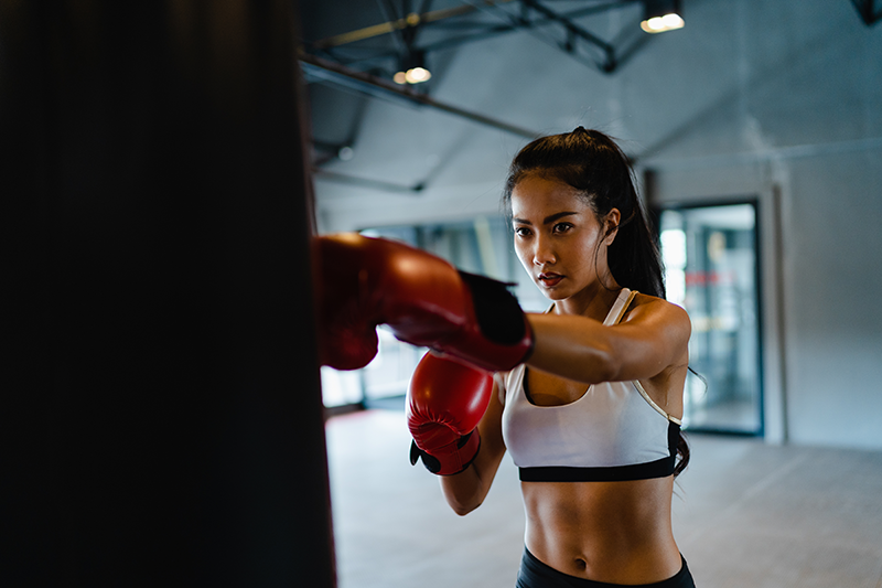 Woman wearing red boxing gloves maintaining intense focus while throwing a straight punch at a heavy bag during a kickboxing workout. Woman wearing red boxing gloves maintaining intense focus while throwing a straight punch at a heavy bag during a kickboxing workout.