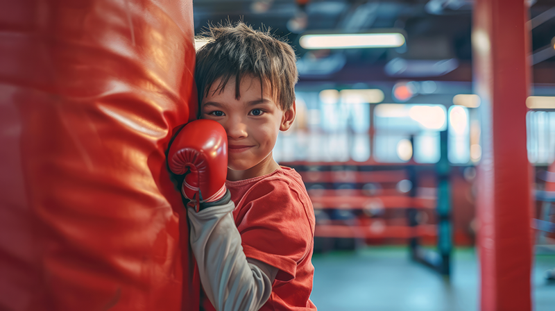 Smiling young boy with boxing gloves resting against a red heavy bag during kids martial arts class