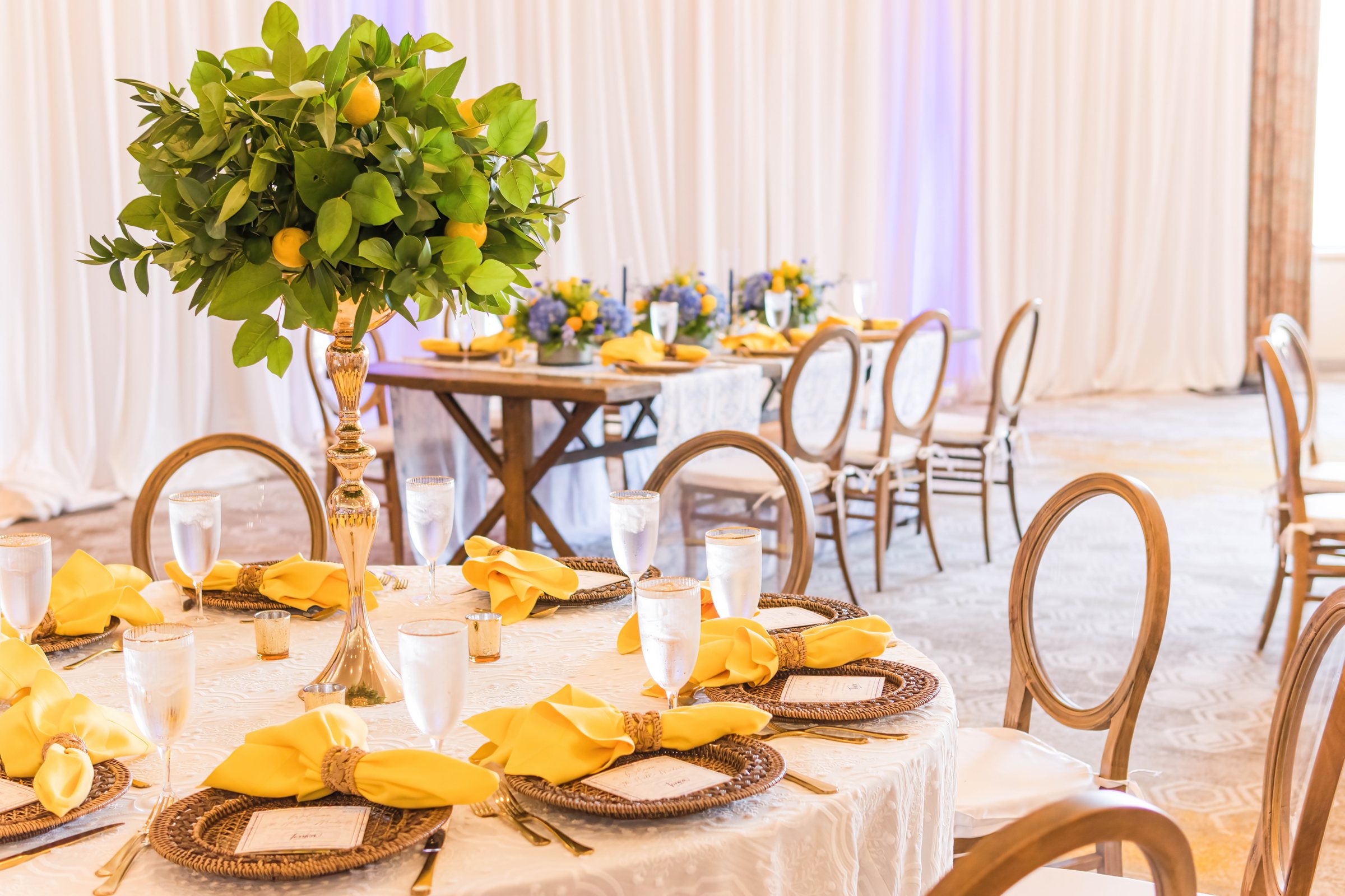 Round table with a tall lemon and greenery centerpiece, yellow napkins, and rattan chargers