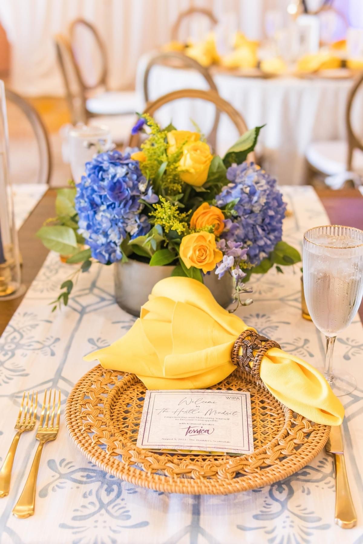 Place setting with WIPA program card, rattan charger, and blue hydrangea arrangement