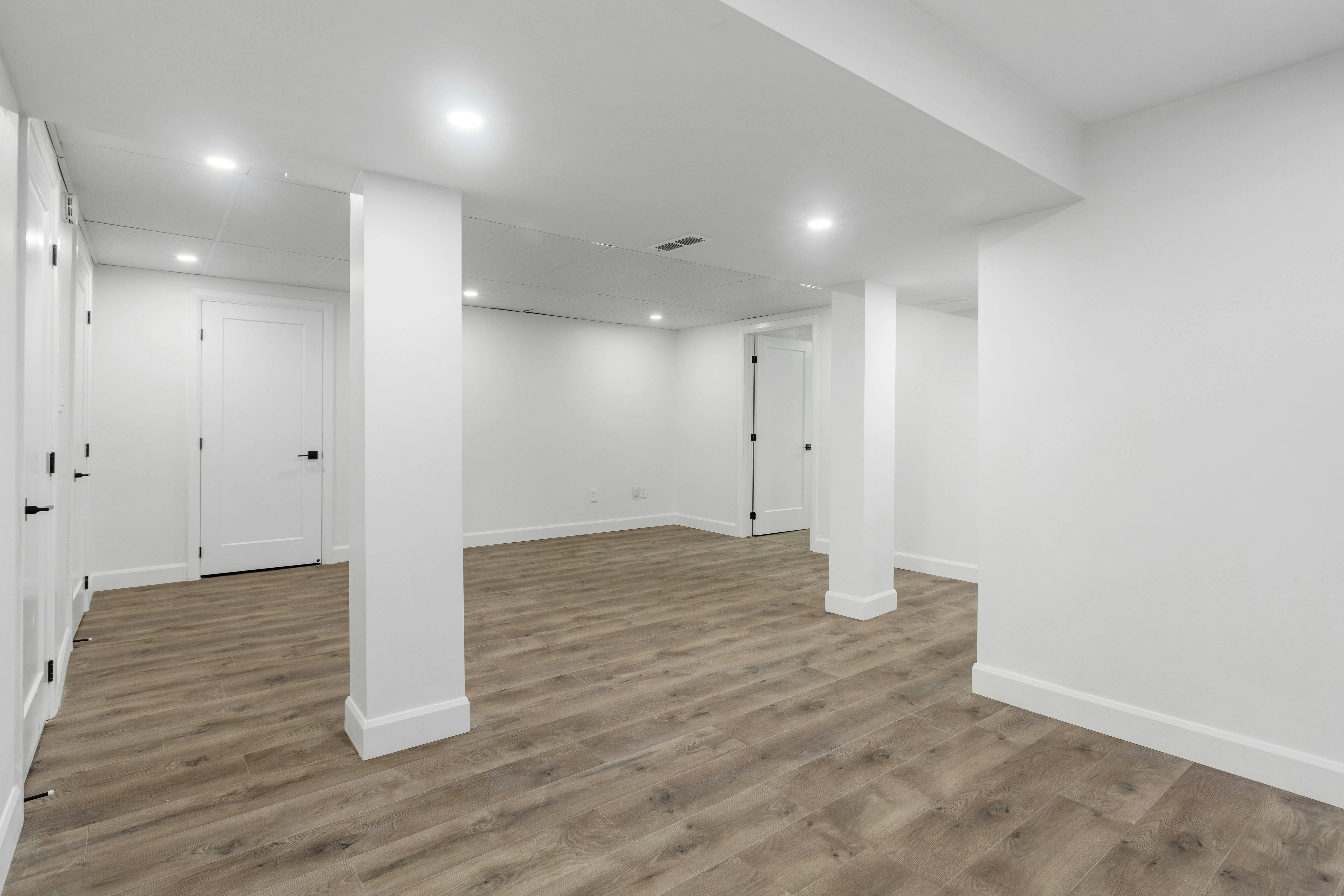 Open basement room with white doors, square columns, recessed lights, and wood-look flooring