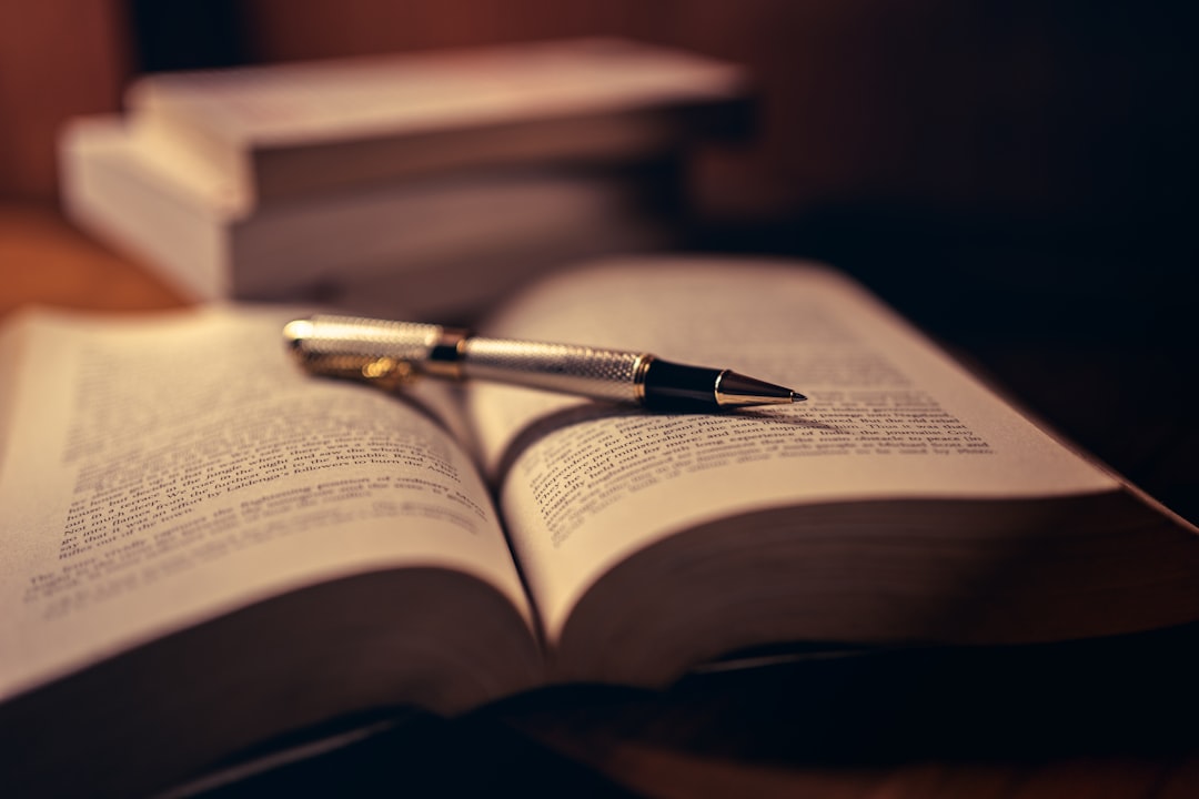 A slim metal pen lying across the pages of an open book, with a stack of closed books blurred in the warm background.