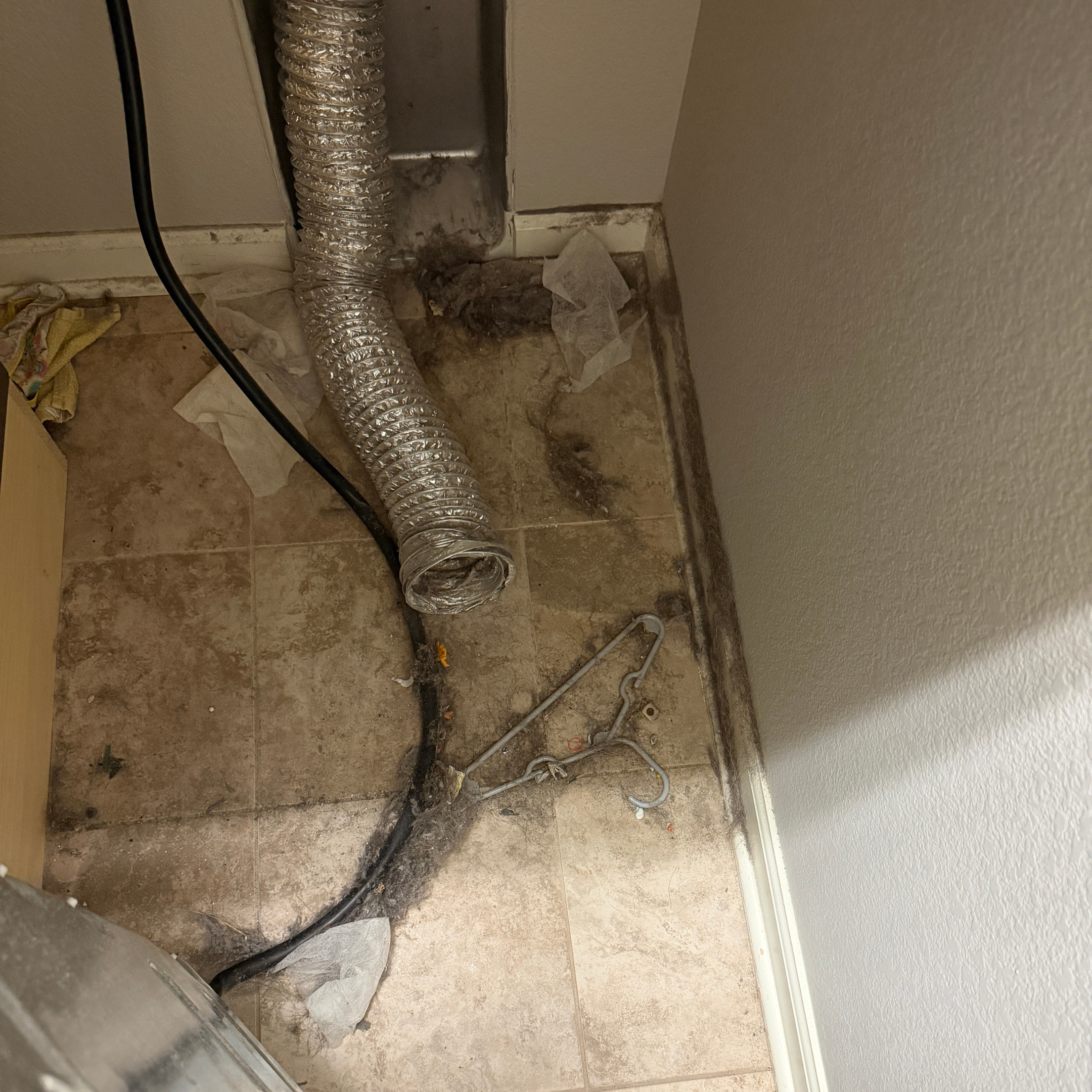 Image is of a dirty dryer vent hose and lint build up behind the dryer. There is also a hanger, dryer sheets and lint on the floor