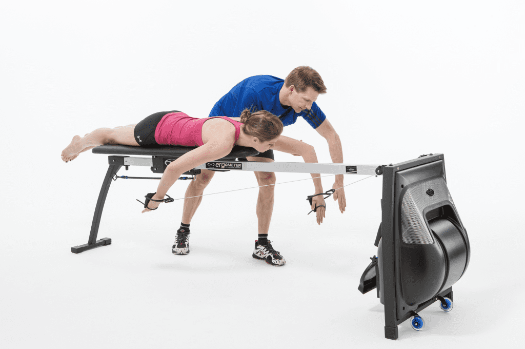 A sports performance coach providing personalized, hands-on feedback to a female athlete analyzing catch and pull mechanics on a specialized swim and rowing ergometer bench.