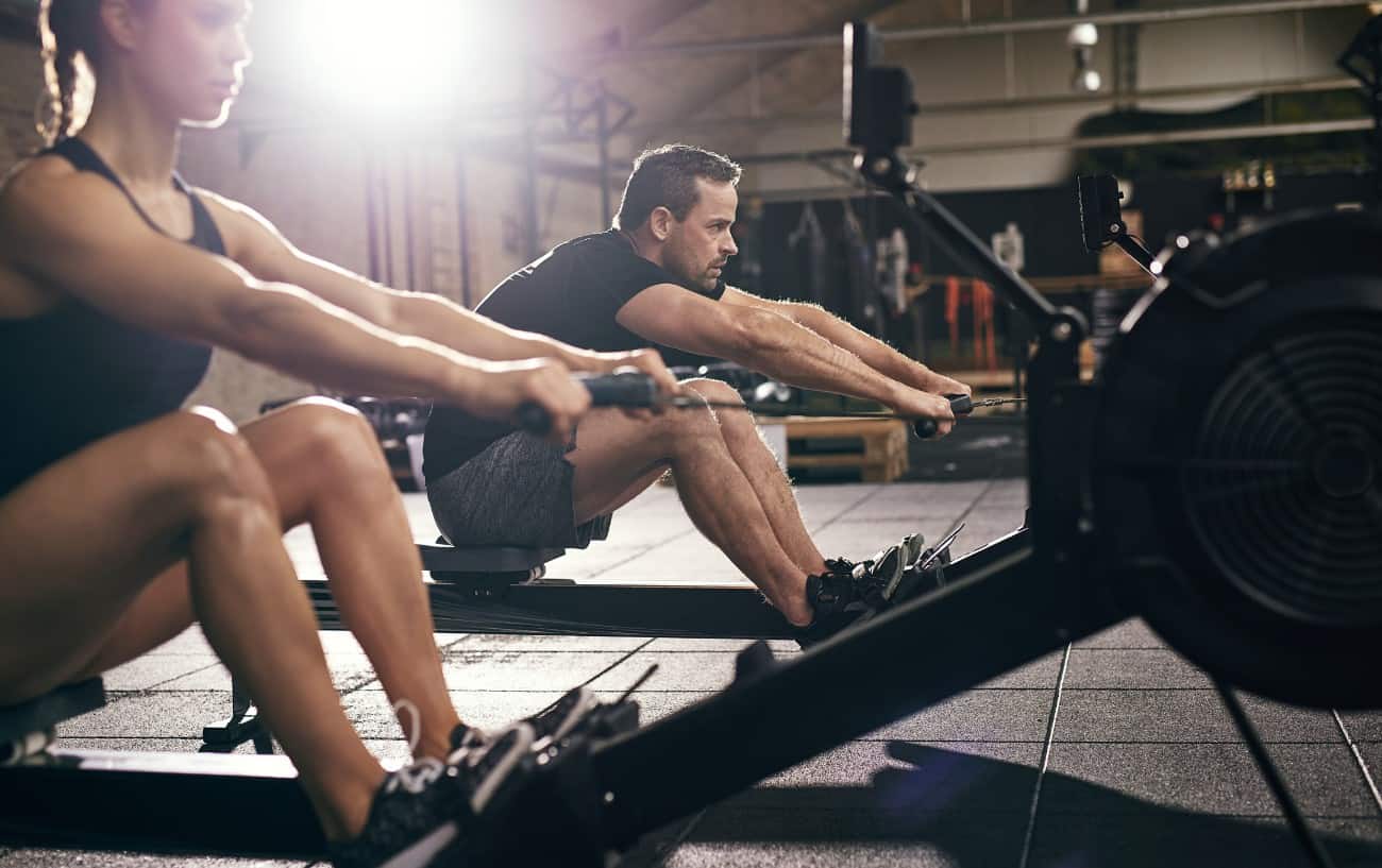 A a man and woman carefully practicing proper rowing technique on indoor rowers in a gym. A a man and woman carefully practicing proper rowing technique on indoor rowers in a gym.
