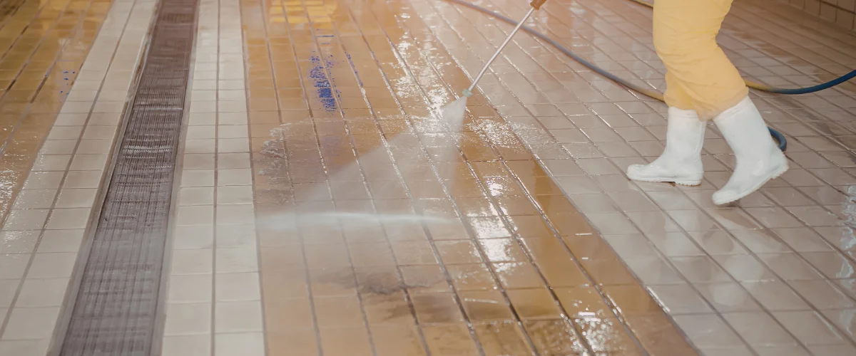 Industrial worker pressure washing tiled floor in facility.