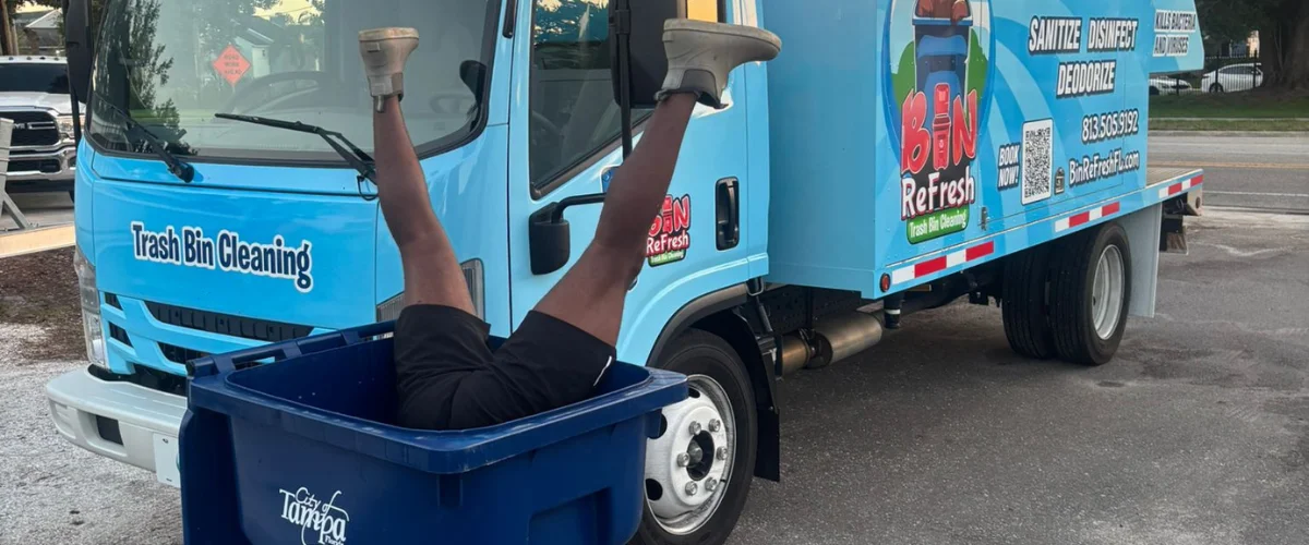 A person's legs upside down in a blue trash bin in front of a Bin Refresh trash bin cleaning truck.