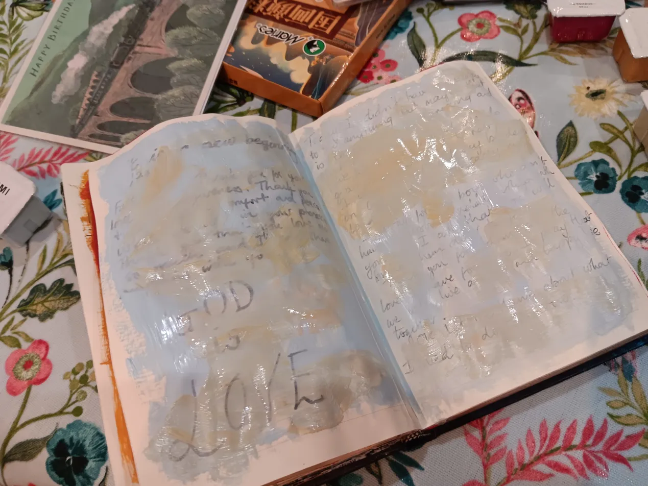 An open art journal with handwritten grief entries painted over in soft blue and cream watercolour washes, with the words "GOD" and "LOVE" visible beneath the paint, resting on a floral tablecloth.