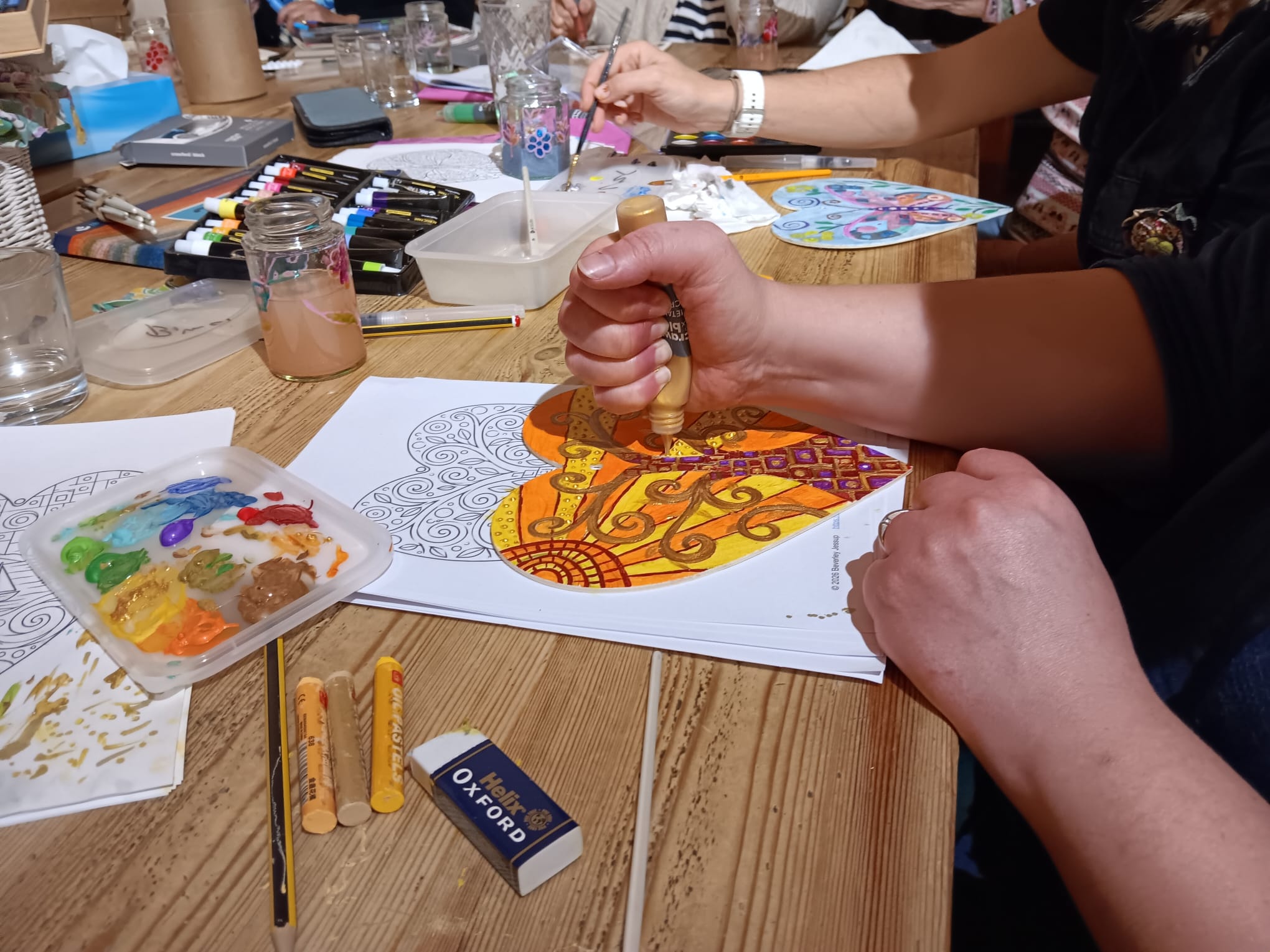 Hands in a craft workshop applying gold paint to a decorative paper heart on a wooden table with paint palettes and art supplies