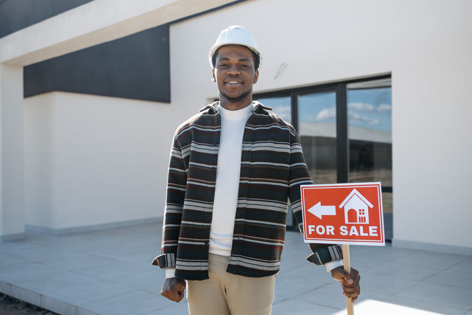 A real estate agent holding a For Sale sign in front of a modern home — illustrative stock photo