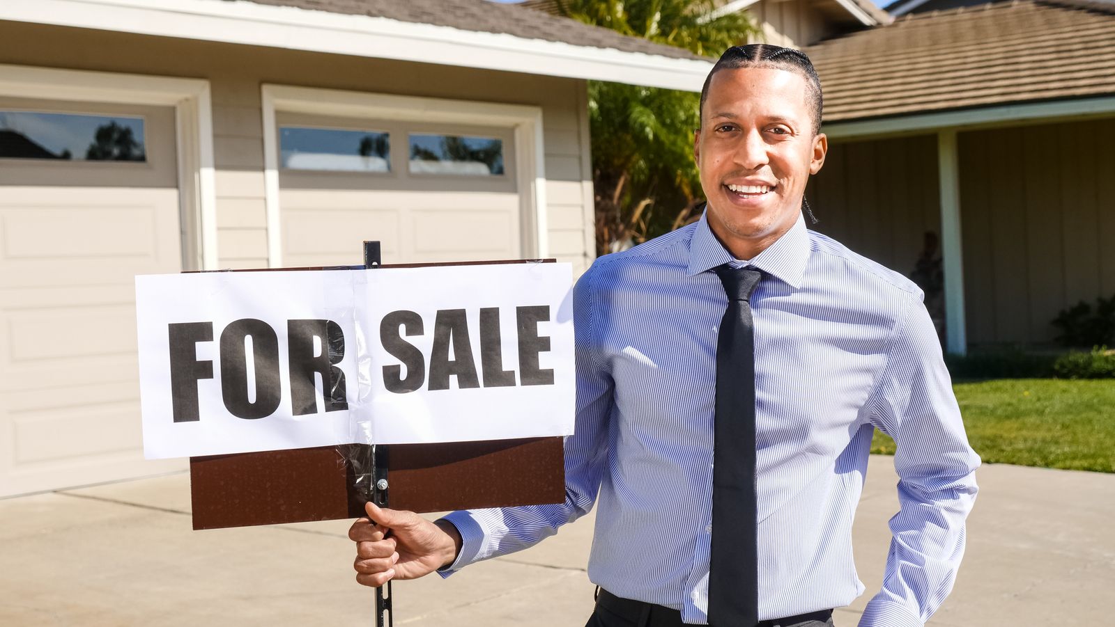 A real estate agent holding a For Sale sign in front of a home — illustrative stock photo