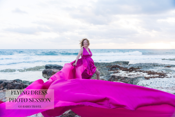 Go Babes traveler wearing a flowing pink flying dress while standing on rocks beside the Caribbean Sea in Belize