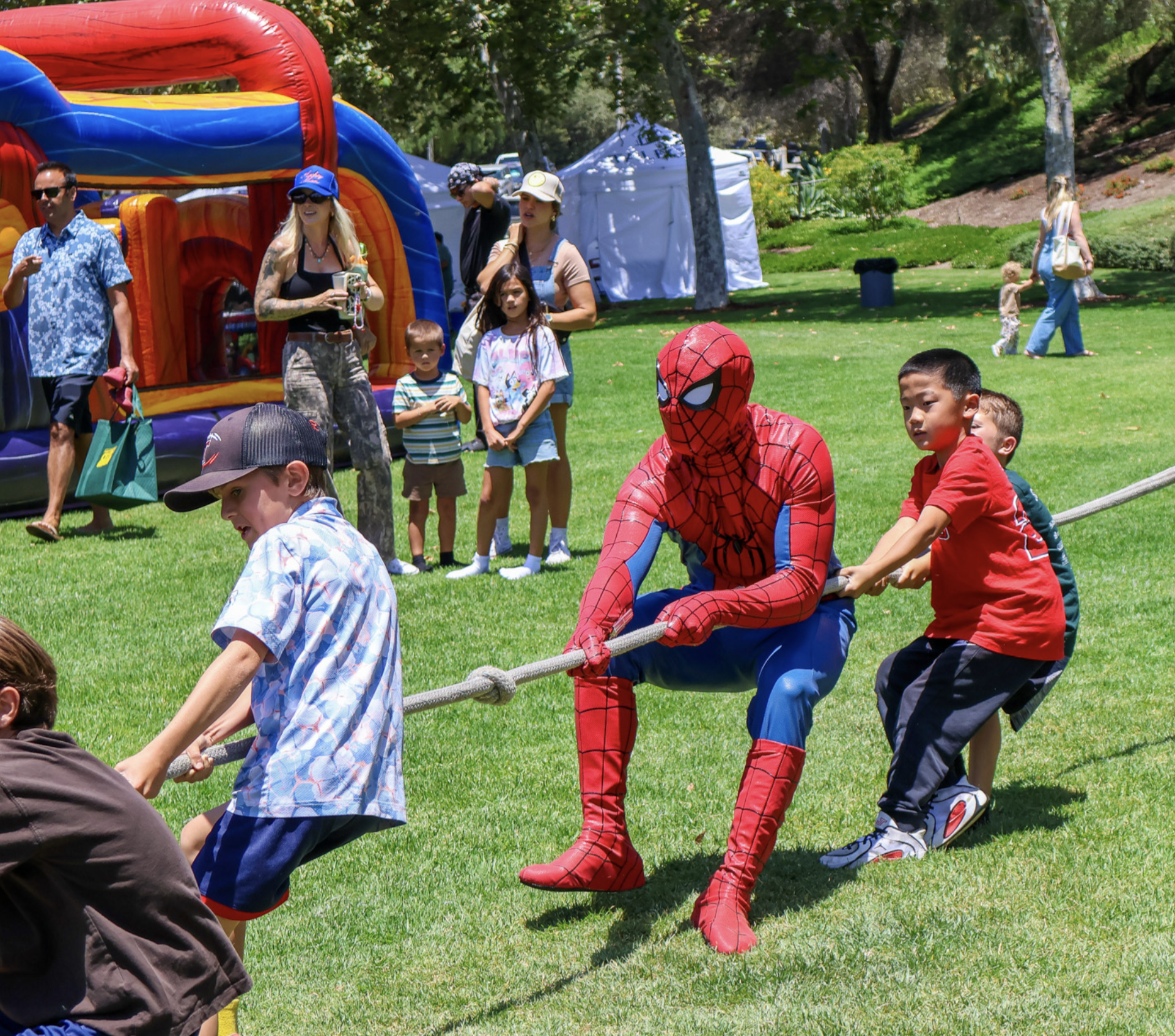 Spider-Man joining children for a group rope game at an outdoor celebration with an inflatable nearby